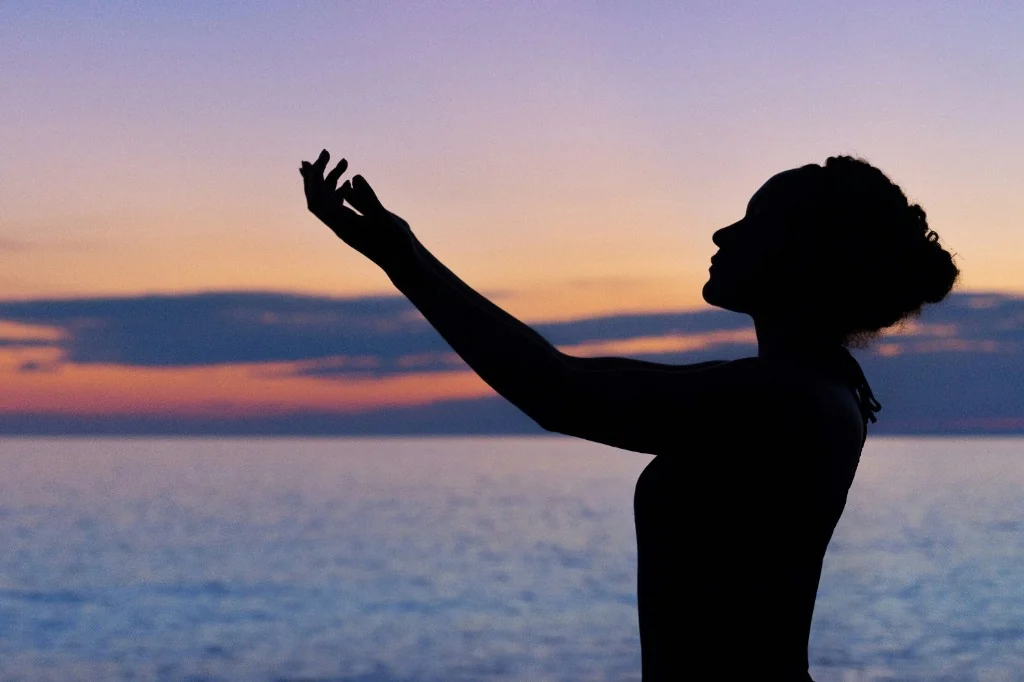 Silhouette of a person with arms raised at sunset over the ocean — a gesture of openness and reflection.